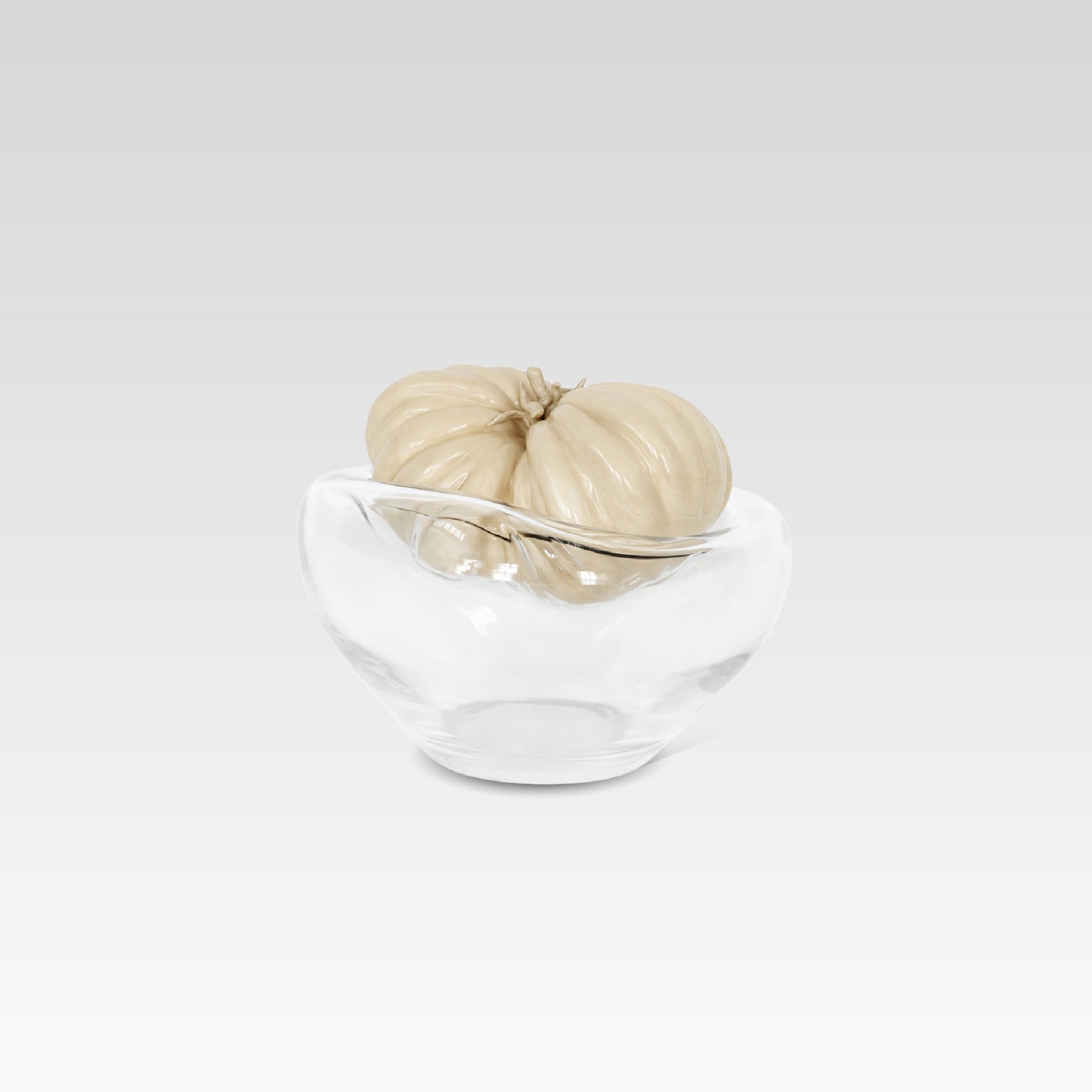 A clear crystal glass vessel with a sculptural, ribbed beige ceramic object resembling a tomato, photographed on a light gray studio background