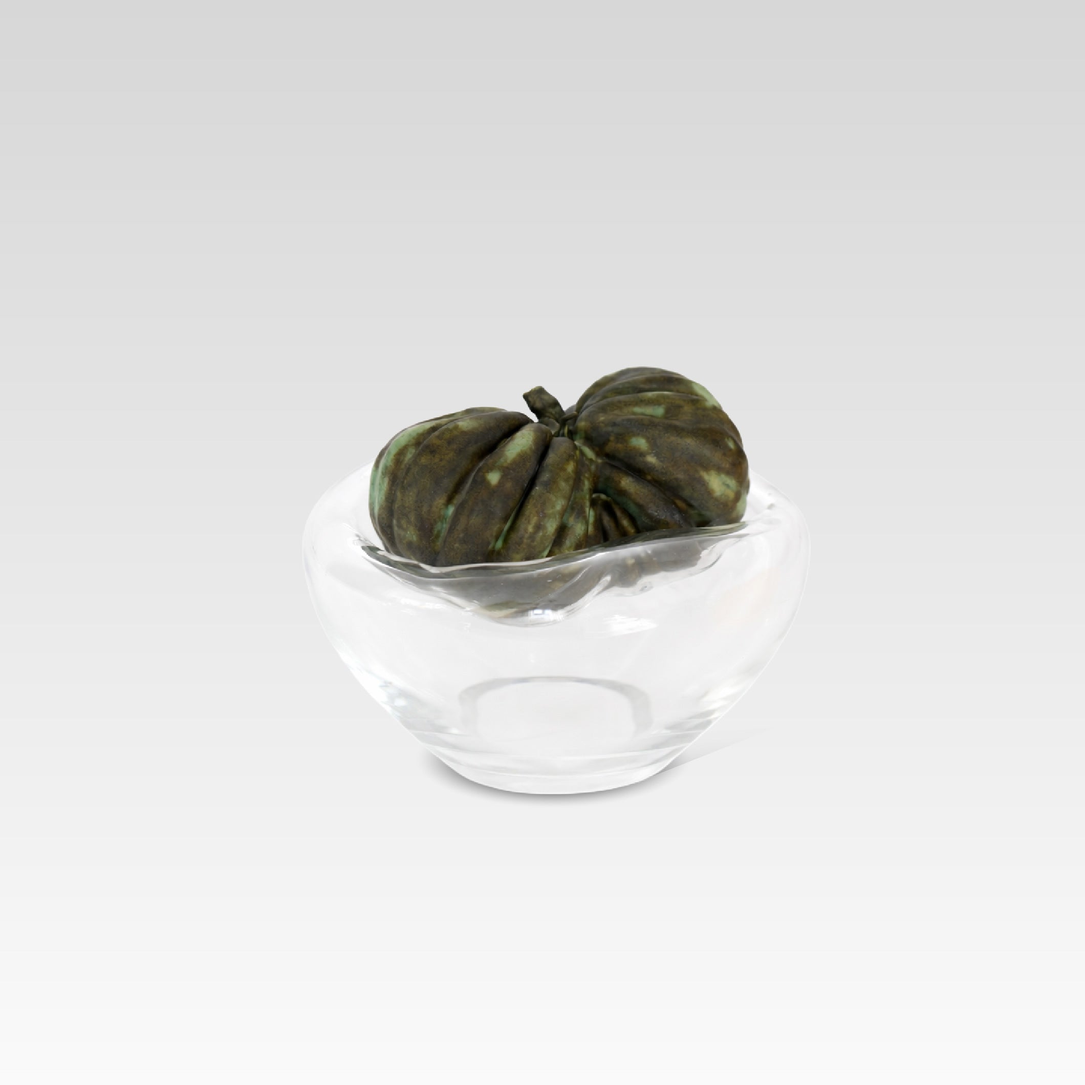A clear crystal glass vessel with a sculptural, ribbed dark brown and organic sculptural ceramic object resembling a tomato, photographed on a light gray studio background