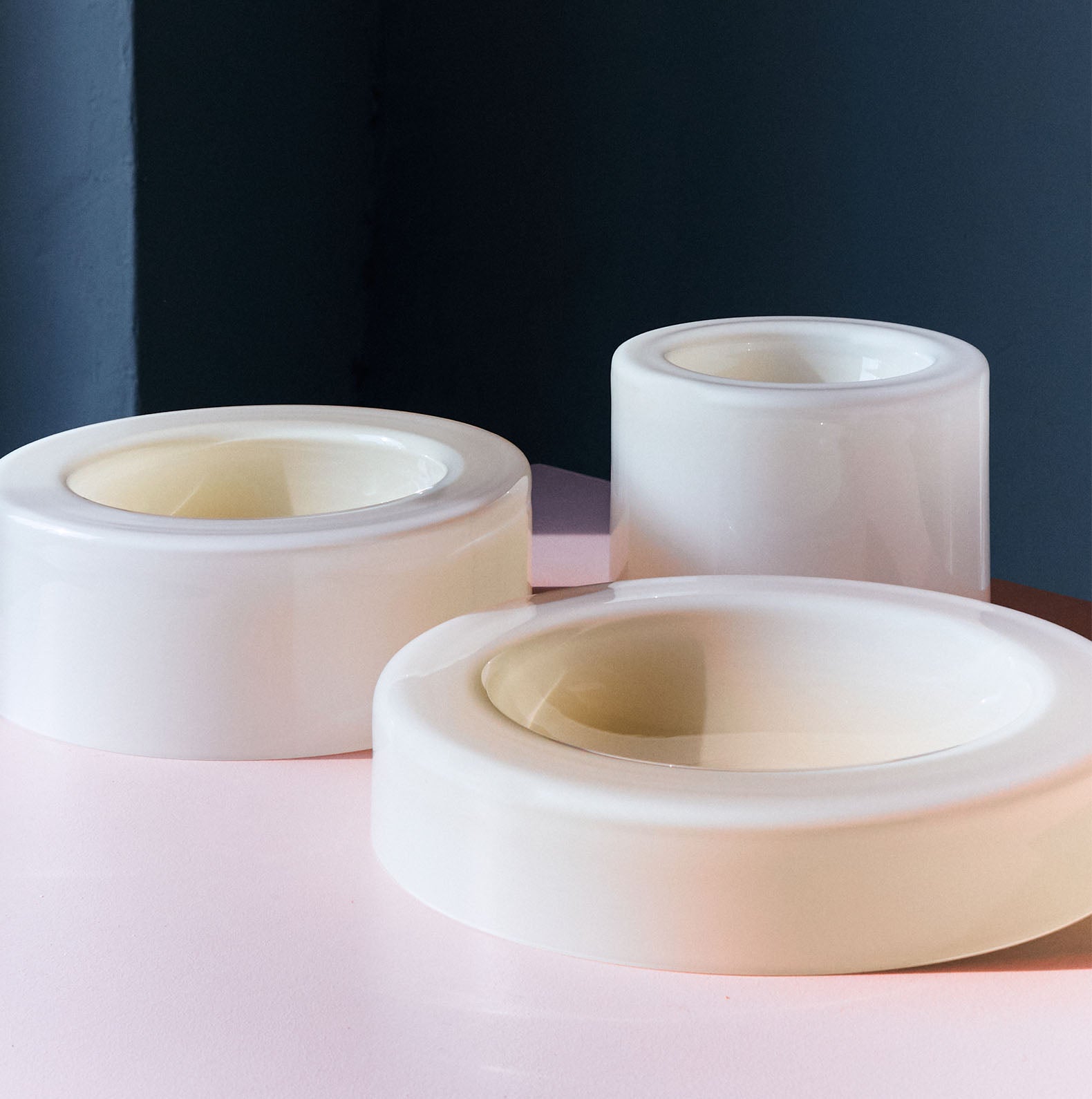 Three smooth cream-white bowls, arranged beautifully on top of a cream pink table, each bowl having different heights and widths, a dark wall in the background. 