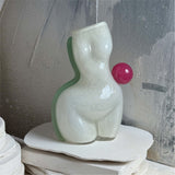 White futura air vase, shaped as a womans figure made of glass with a light blue/green stripe on the left and a pink ball on the right waist, positioned on a textured background