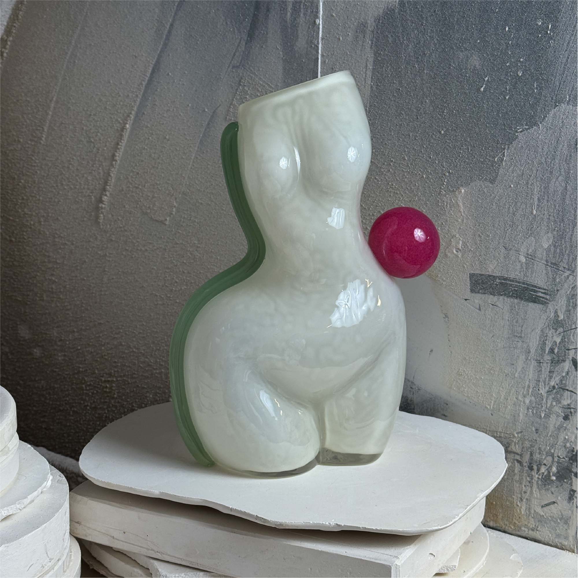 White futura air vase, shaped as a womans figure made of glass with a light blue/green stripe on the left and a pink ball on the right waist, positioned on a textured background