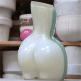 White futura air vase, shaped as a womans figure from behind, made of glass, with a light blue/green stripe on the right and a pink ball on the right waist, positioned in a workshop setting
