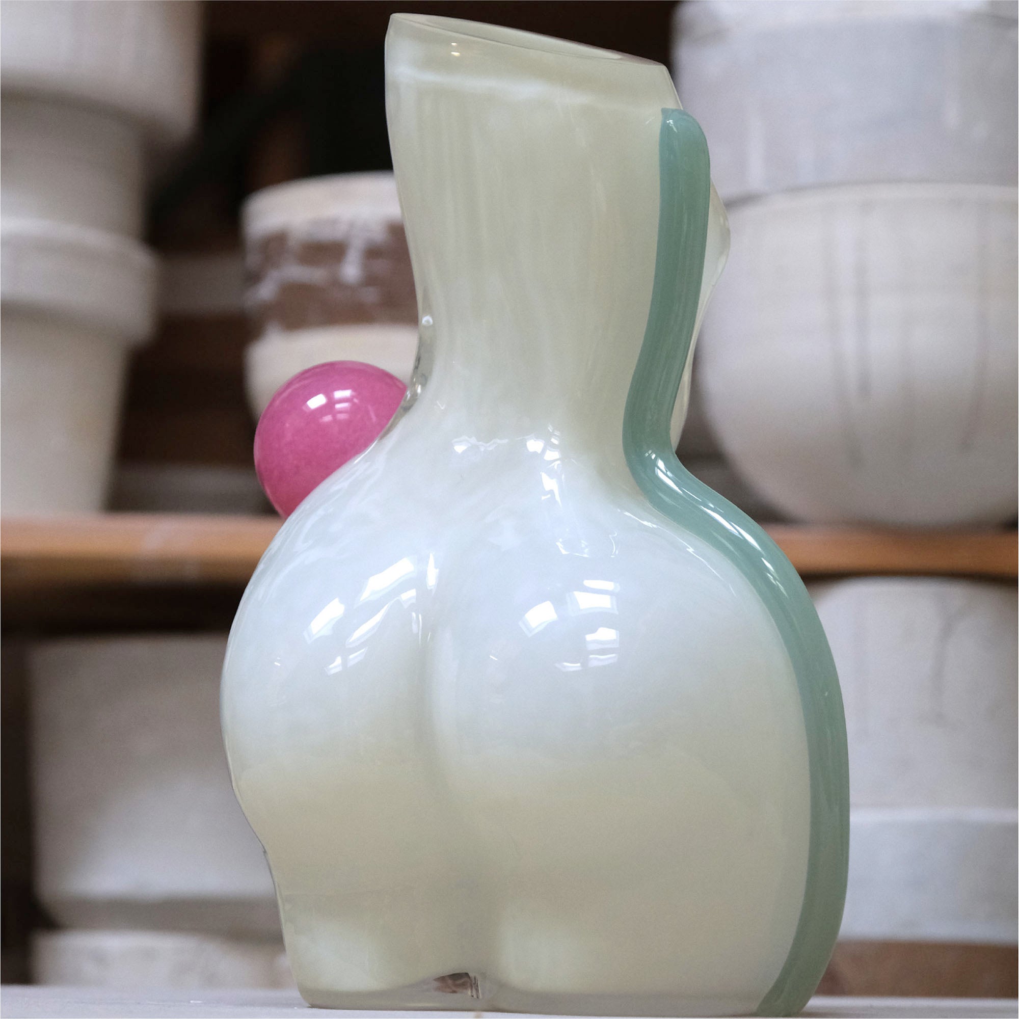 White futura air vase, shaped as a womans figure from behind, made of glass, with a light blue/green stripe on the right and a pink ball on the right waist, positioned in a workshop setting