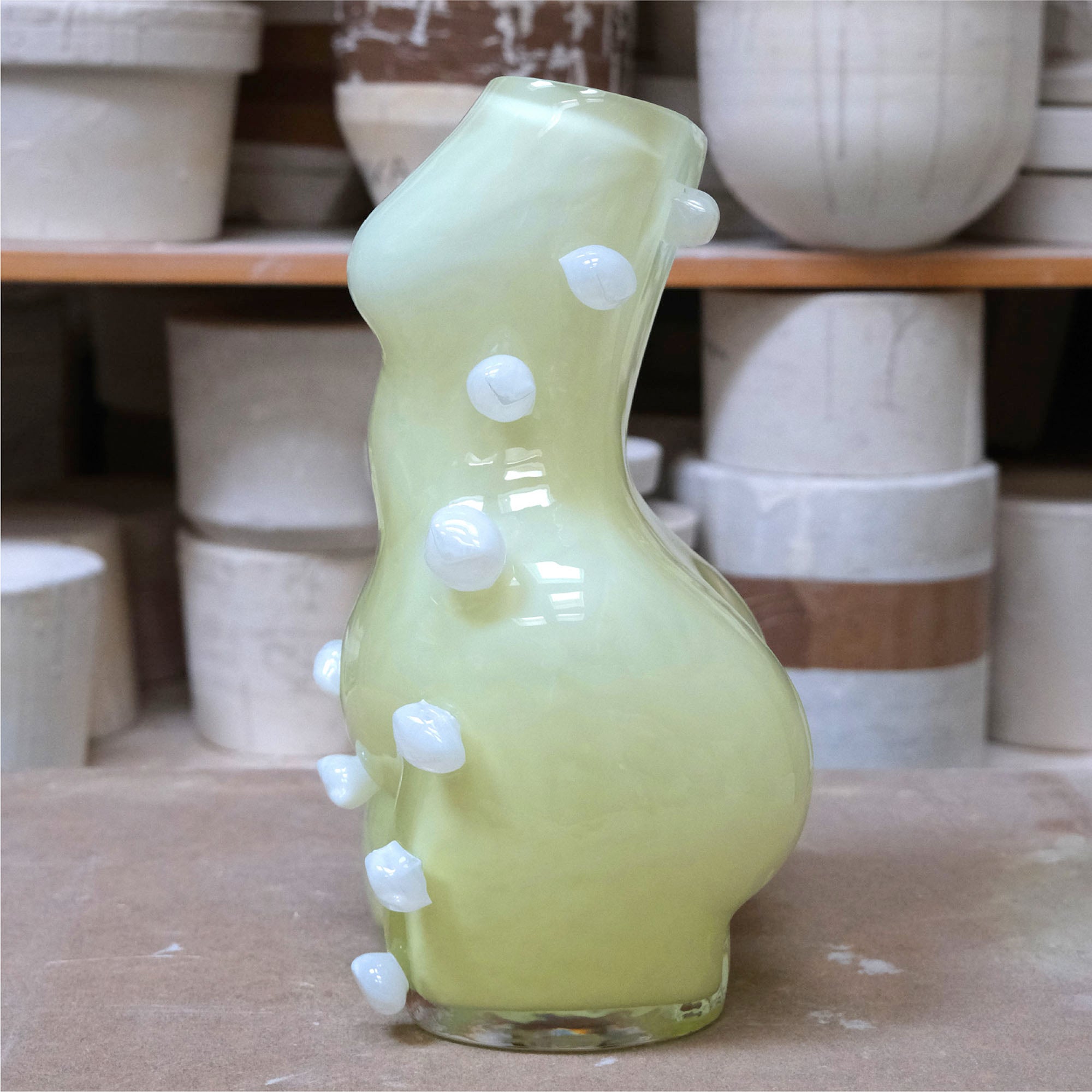 Limoncello coloured glass vase with white blobs on a wooden surface with ceramic pots in the background