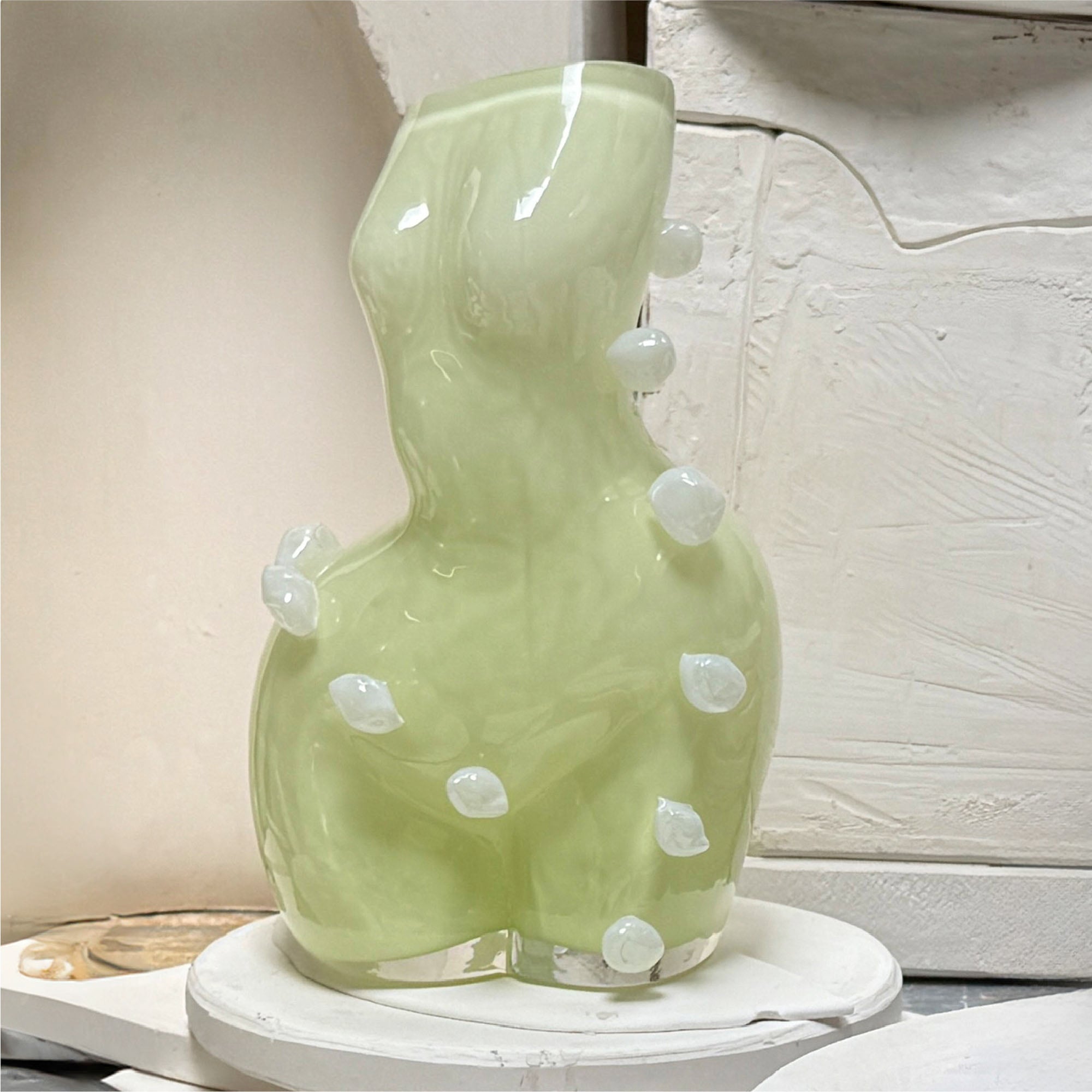 Futura cloud limoncello coloured womans figure, glass vase, with white blob dots on a white surface in a workshop context.