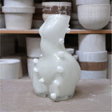 Glass sculpture with white knobs on a wooden surface, with shelves of ceramic pots in the background.