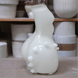 White ceramic vase with unique womans figure shape on a wooden surface, with shelves of vases in the background.