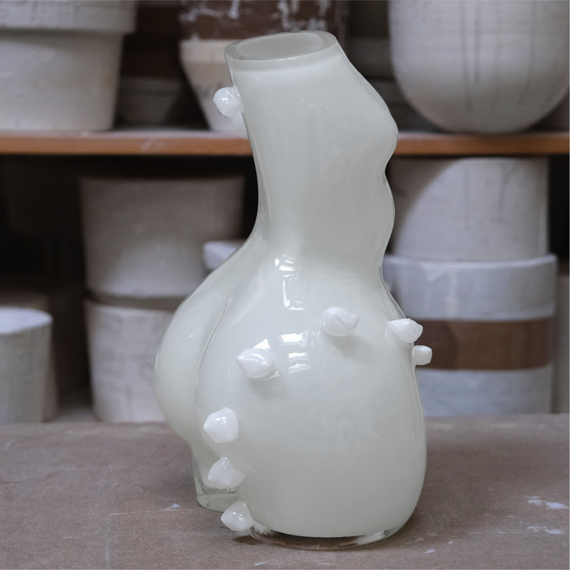 White ceramic vase with unique womans figure shape on a wooden surface, with shelves of vases in the background.