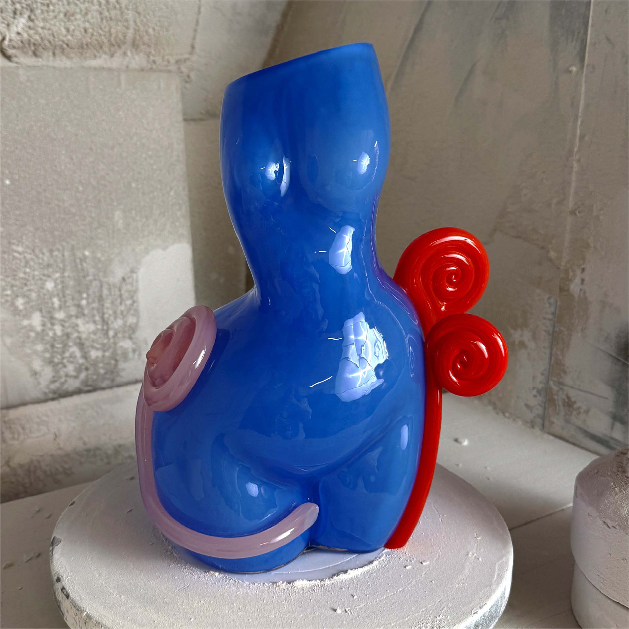 Blue and red sculpture on a white pedestal against a textured wall.