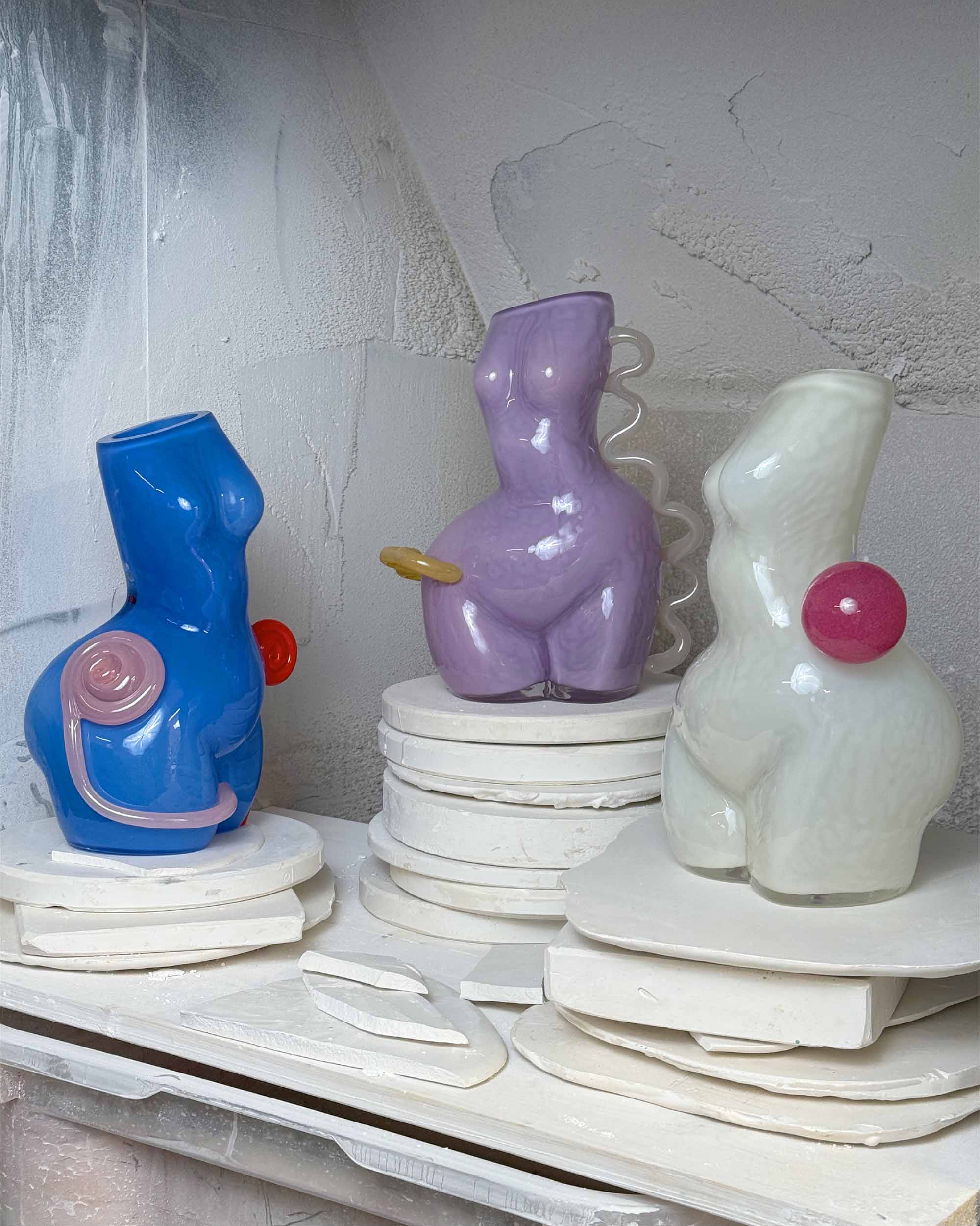Three glass women figure sculptures on a white surface with a textured wall background