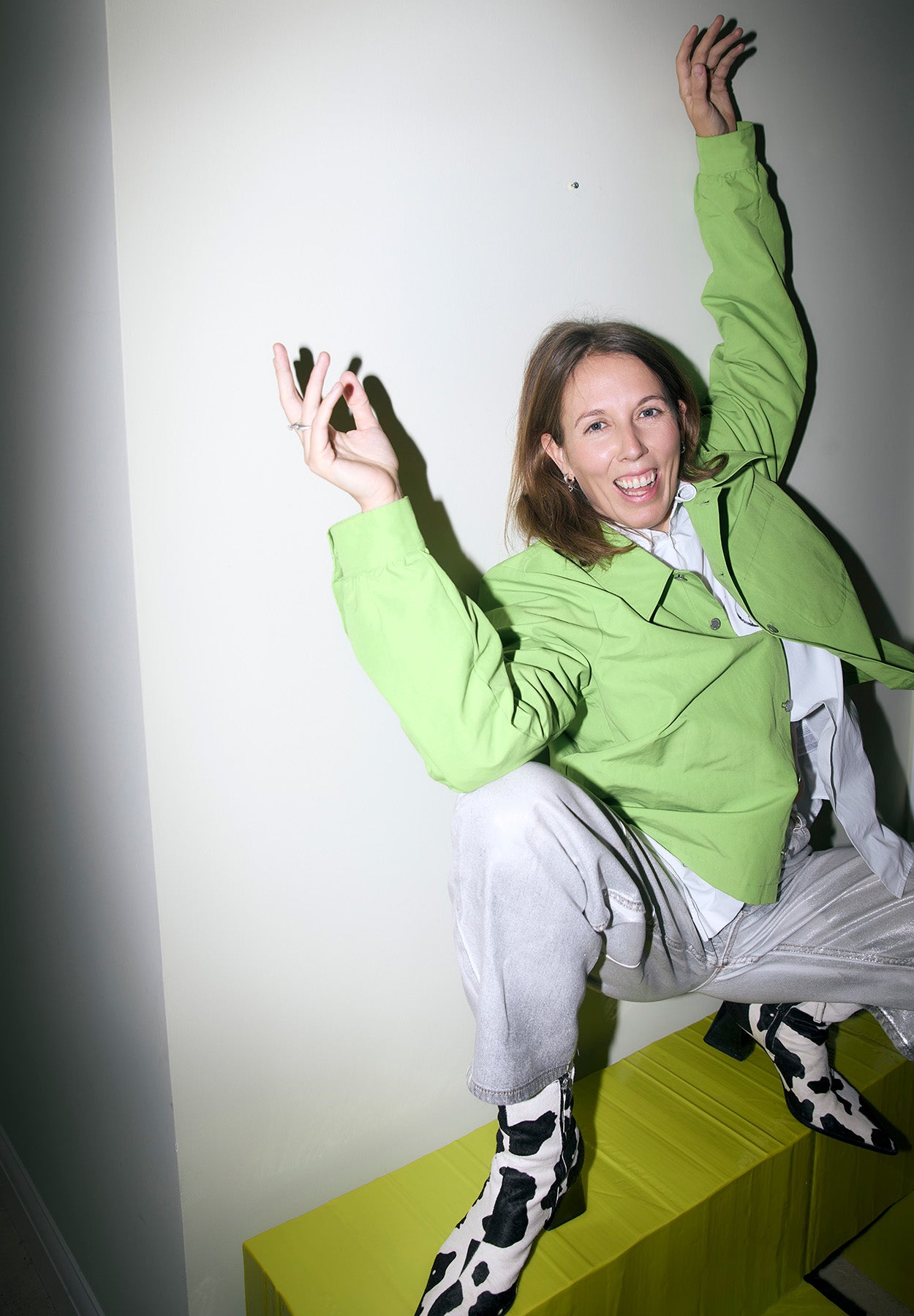 Kathrin, founder of Ursula Futura, mid-jump on a yellow-green bench in a green jacket and cow-print boots.