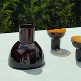 A Brown Plash Carafe with two Brown Plash Glasses are positioned on a light textured table with greenery in the background.
