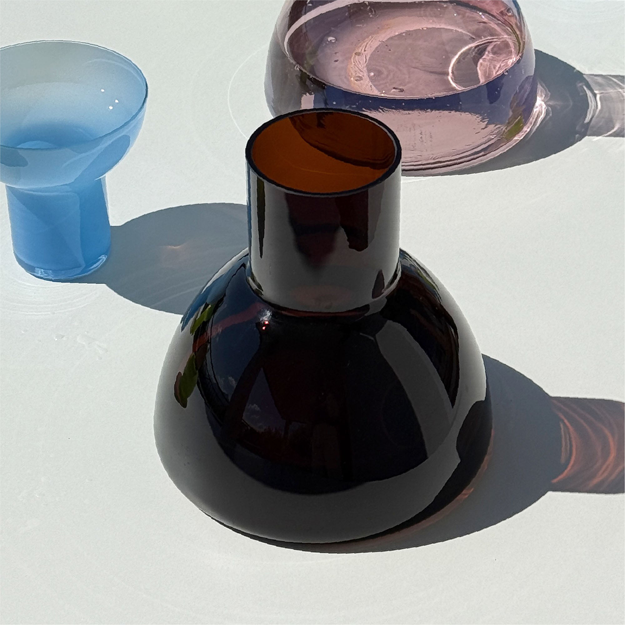 Brown Carafe withsunlight coming from the left creating a shadowdrop toward the right. Another carafe is visible just behing as well as a plash glass to the left. Positioned on top of a white table.
