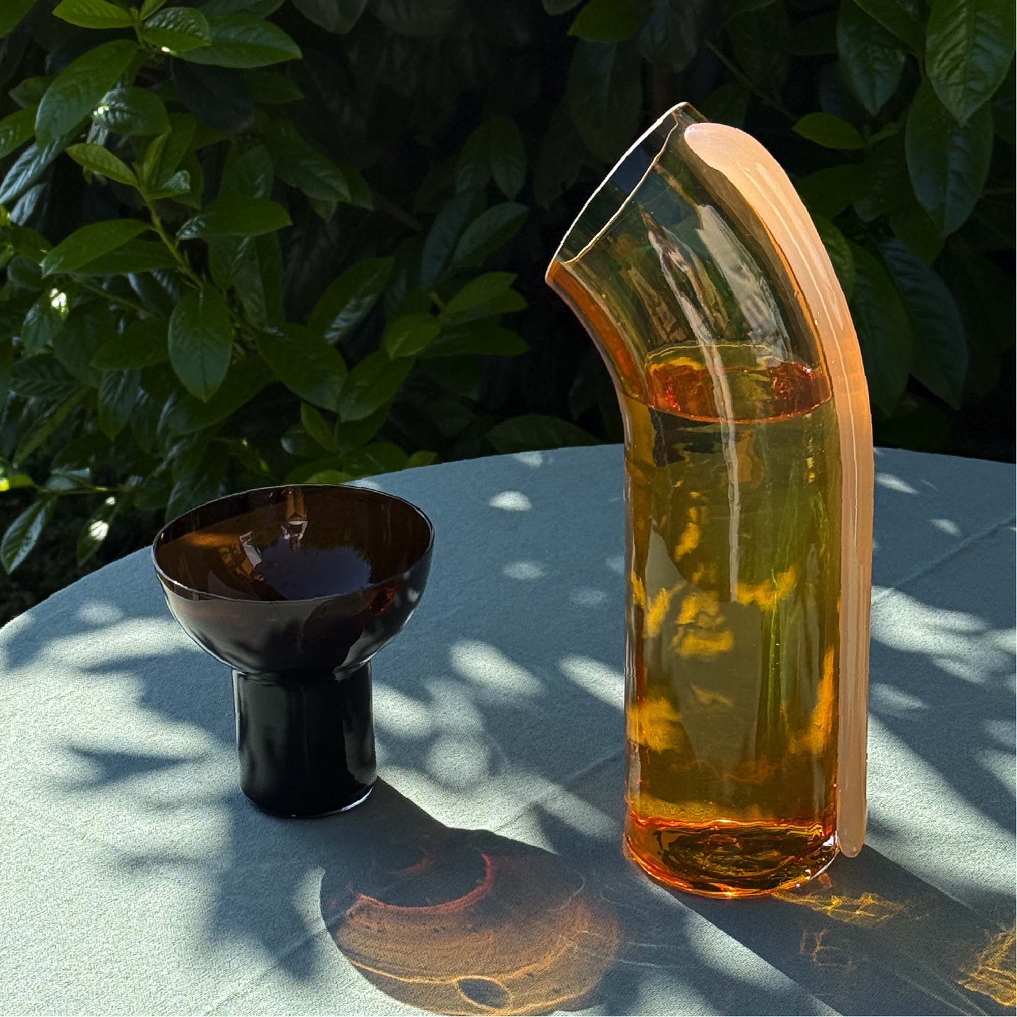 Glass Nessie decanter with a pale apricot stripe on the back positioned on a table in a natural scenery. A dark brown glass is positioned next to the Nessie carafe. 