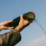 A persons two hands holding the Nessie carafe as water pours out of it. The blue sky is in the background.
