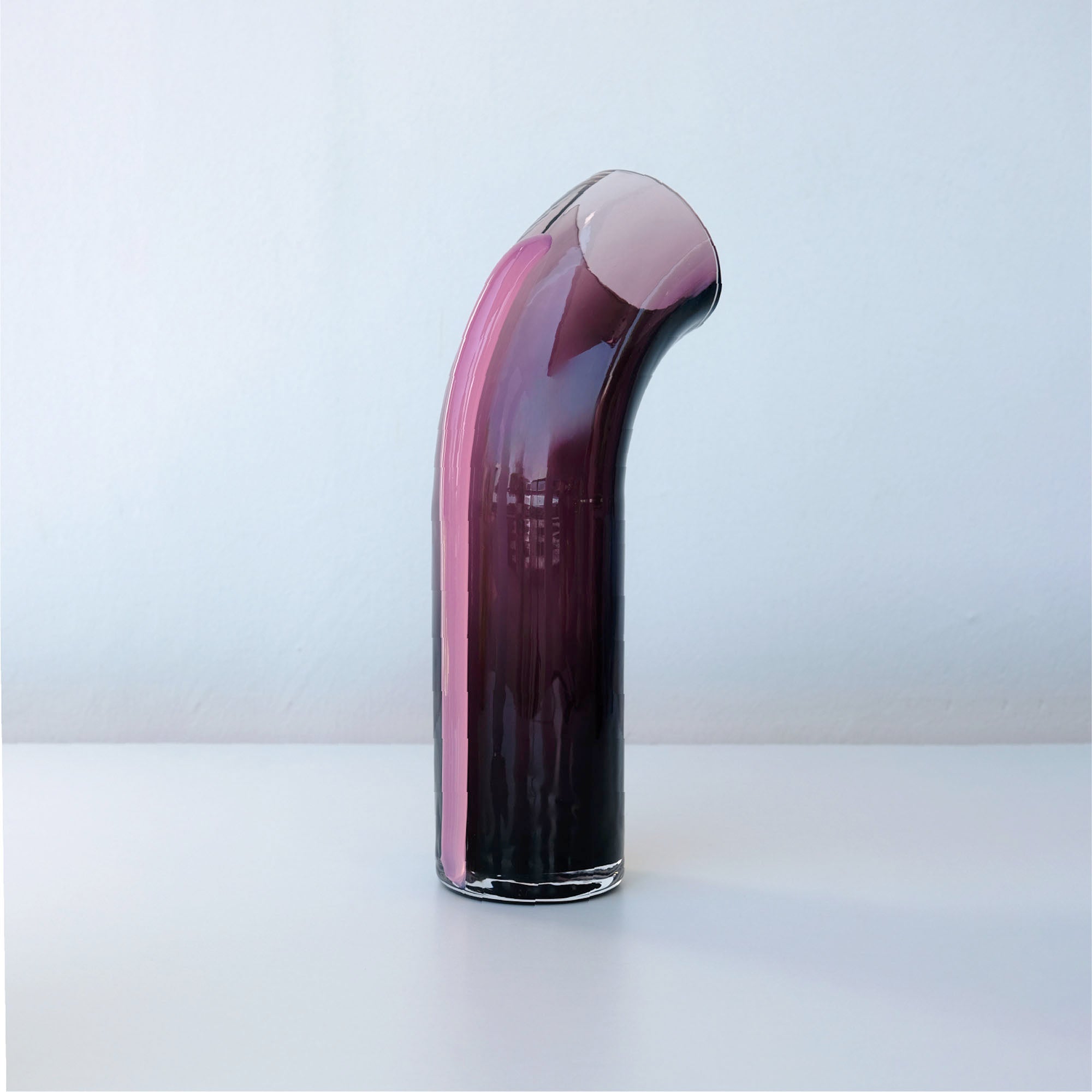 Dark purple glass cylinder with a slight arc on top facing away from the camera, with an opening at the top, a light pink strip of glass connected to the back of the vase, on a white background