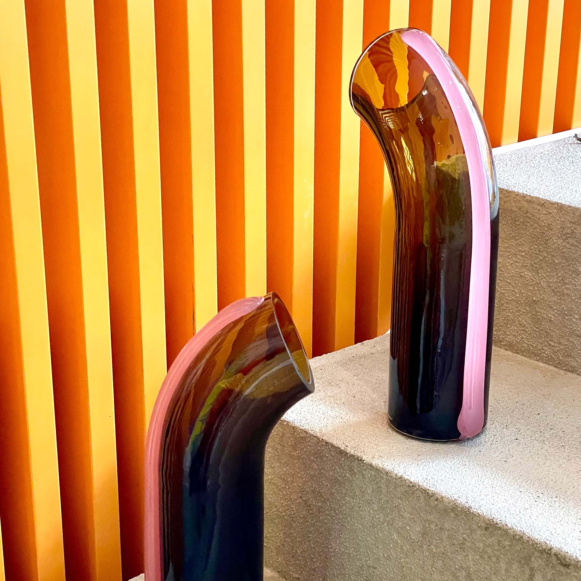 Two Olive-bown Nessie carafes with a pink-purple strip on the back, on a grey concrete stair case against a stripy orange wall.