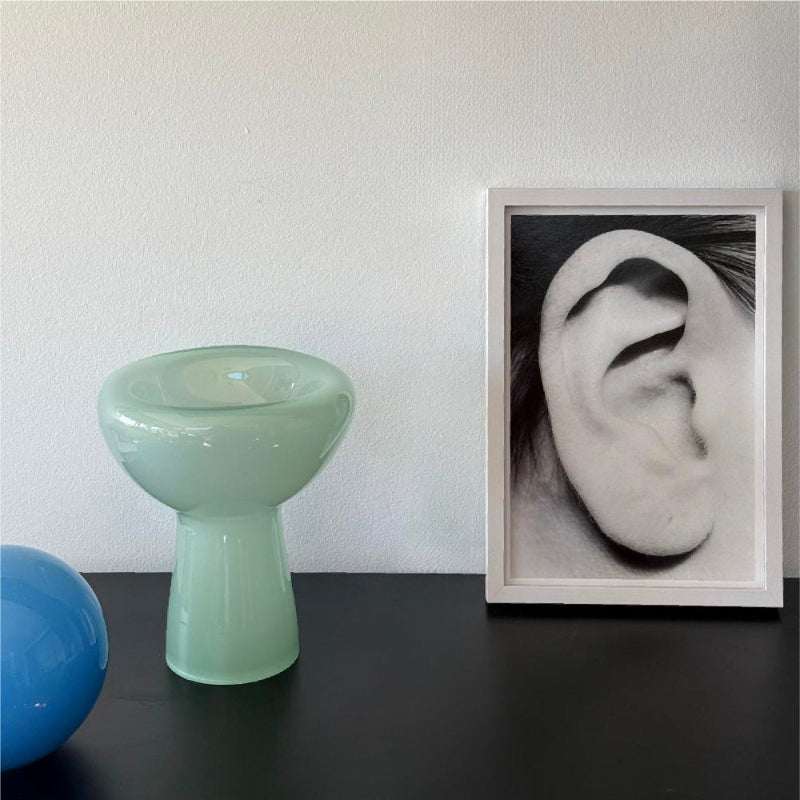 Green vase and blue ball on a dark surface with a framed black and white photo of an ear on a light gray wall.
