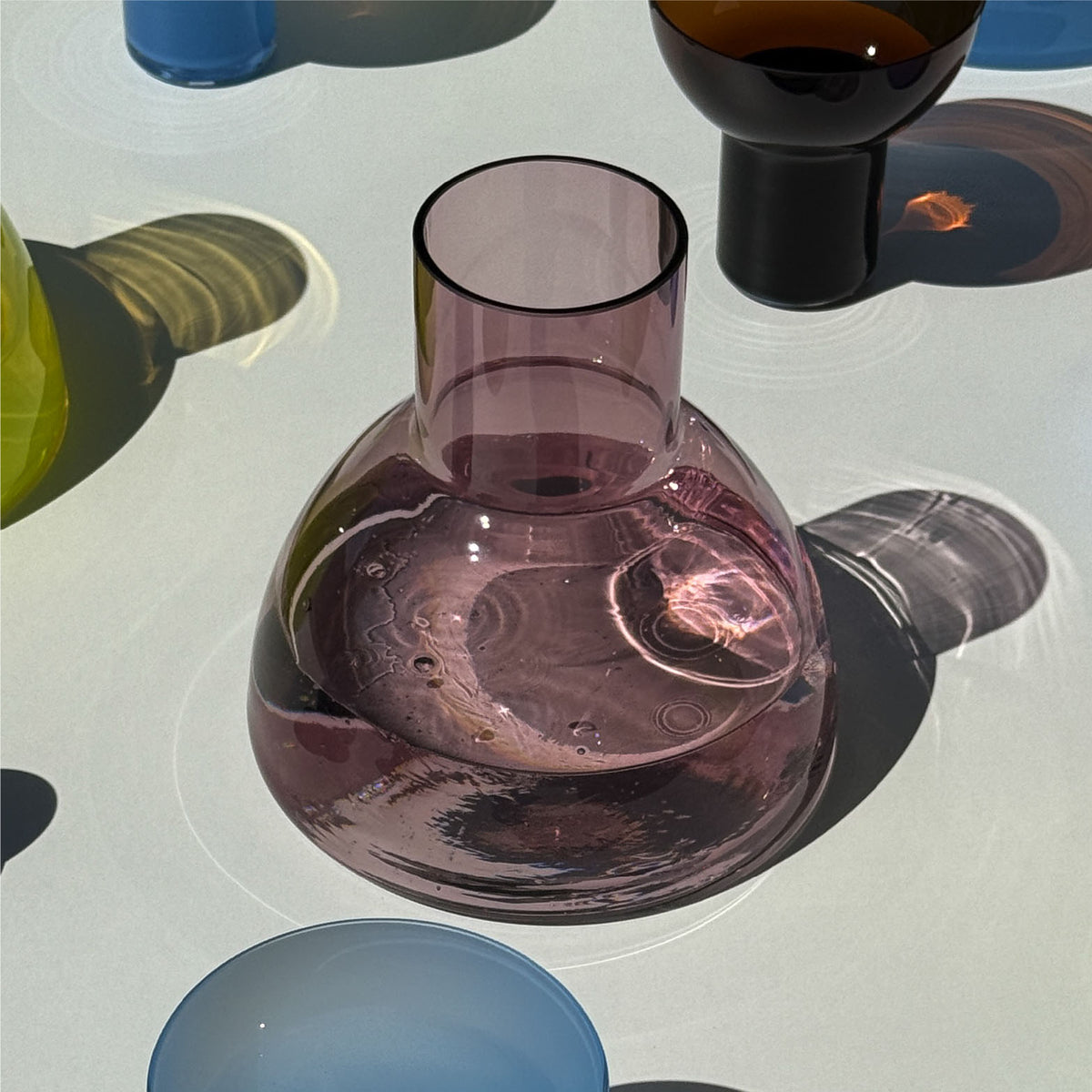 A translucent aubergine glass carafe with a wide rounded base and a short neck, shown on a glossy white surface with colorful glassware in the background.