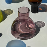 A translucent aubergine glass carafe with a wide rounded base and a short neck, shown on a glossy white surface with colorful glassware in the background.