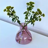 A translucent aubergine coloured plash carafe glass, being used as a vase holding green flower stems with small round yellow-green blooms, set on a white surface against a white wall.