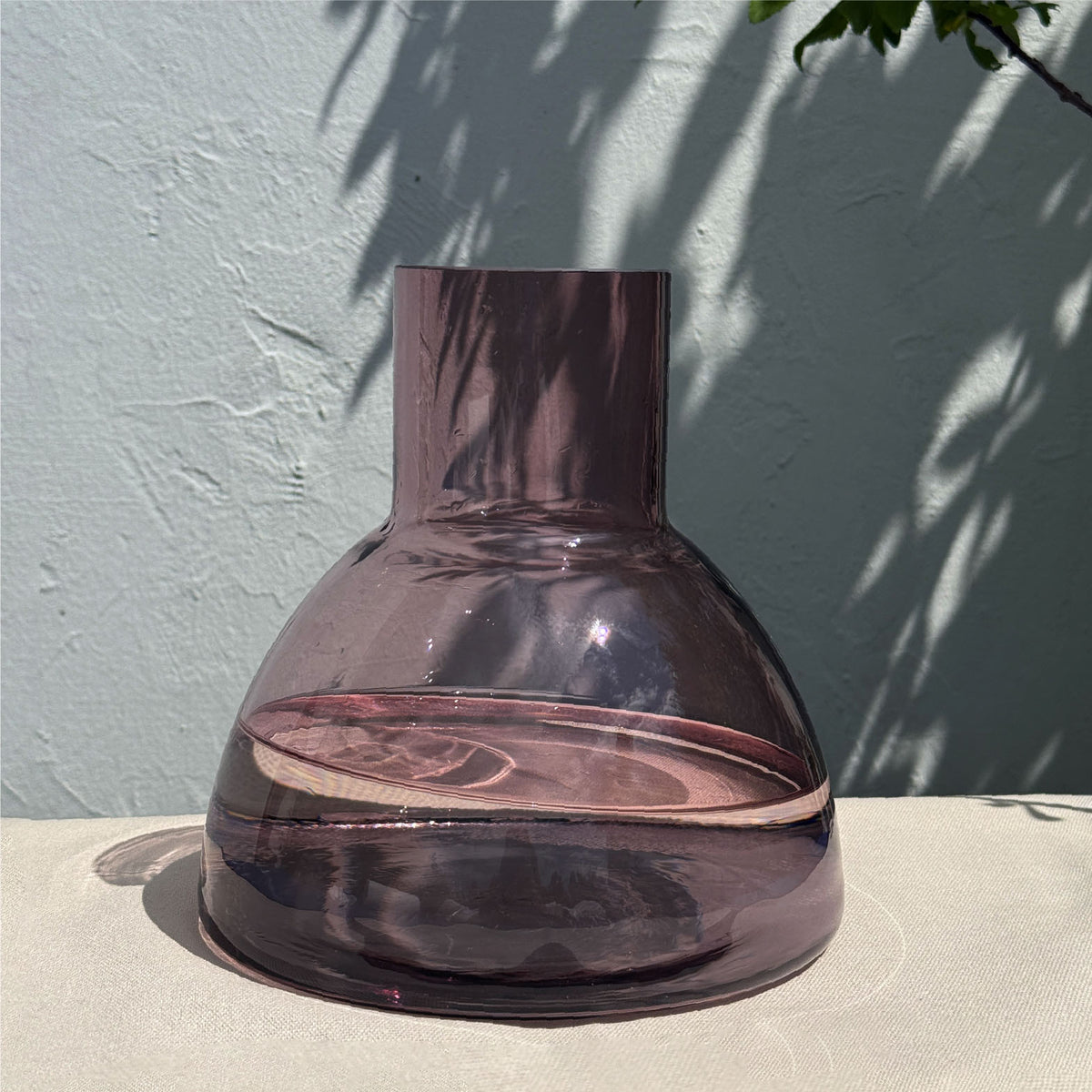 An aubergine coloured plash carafe glass with a wide rounded base and a short cylindrical neck, set on a light beige surface against a textured light blue-gray wall with leaf shadows.