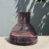 An aubergine coloured plash carafe glass with a wide rounded base and a short cylindrical neck, set on a light beige surface against a textured light blue-gray wall with leaf shadows.