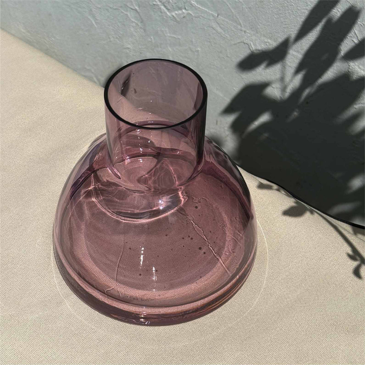 A translucent aubergine glass carafe with a wide, rounded base and a short cylindrical neck, set on a textured beige surface with leaf shadows cast across it.