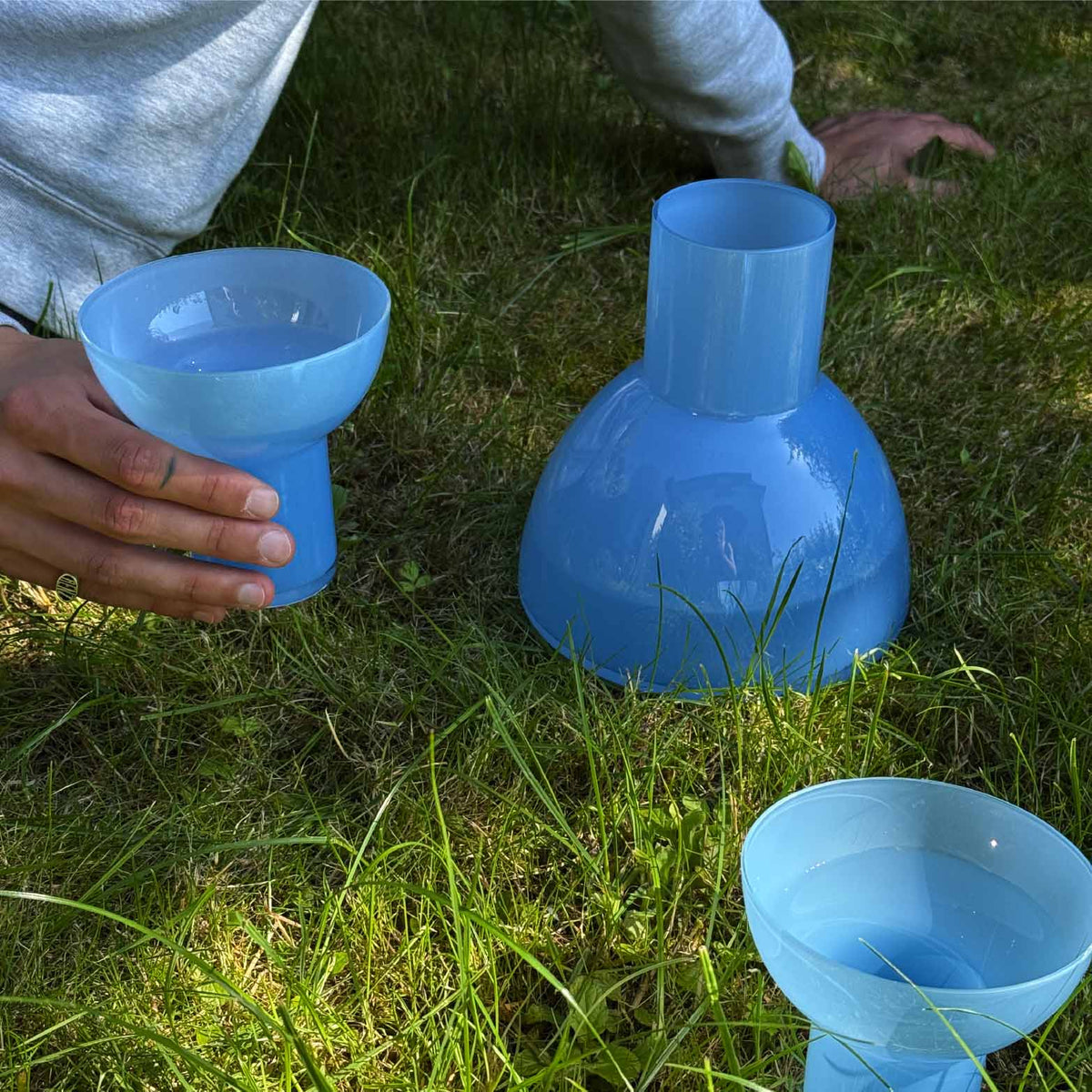 Three light blue glass plash carafes and glasses arranged on green grass outdoors, with a person's hands holding one blue glass and another visible in the foreground.