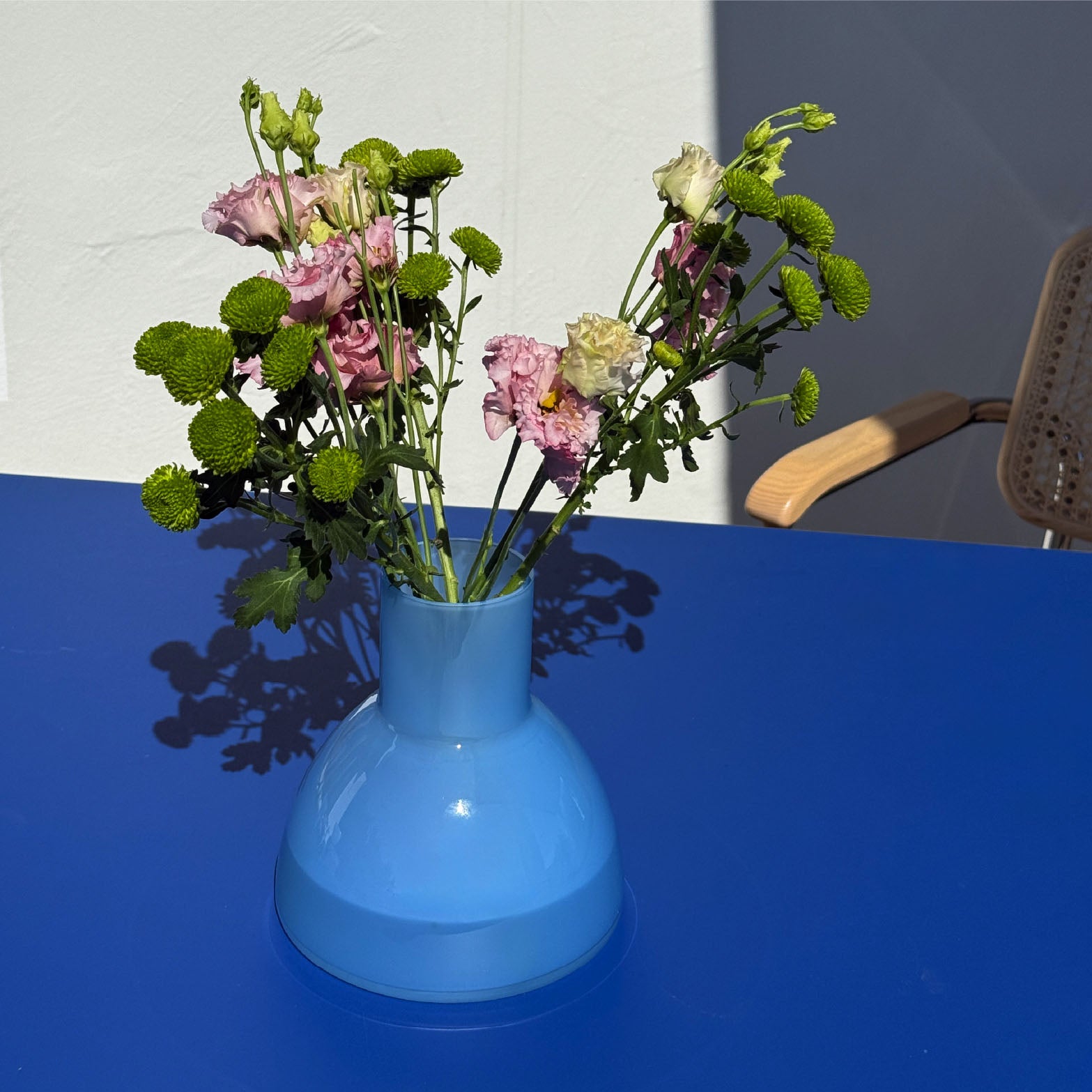 A light blue Plash carafe used as a vase filled with pink and green flowers, placed on a bright blue table against a white wall with a chair partially visible.