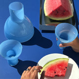 Three light blue glass objects arranged on a dark blue surface, with hands holding and using them near watermelon slices on a plate and tray. A human hand is visible, so this is Lifestyle rather than Product/Packaging.