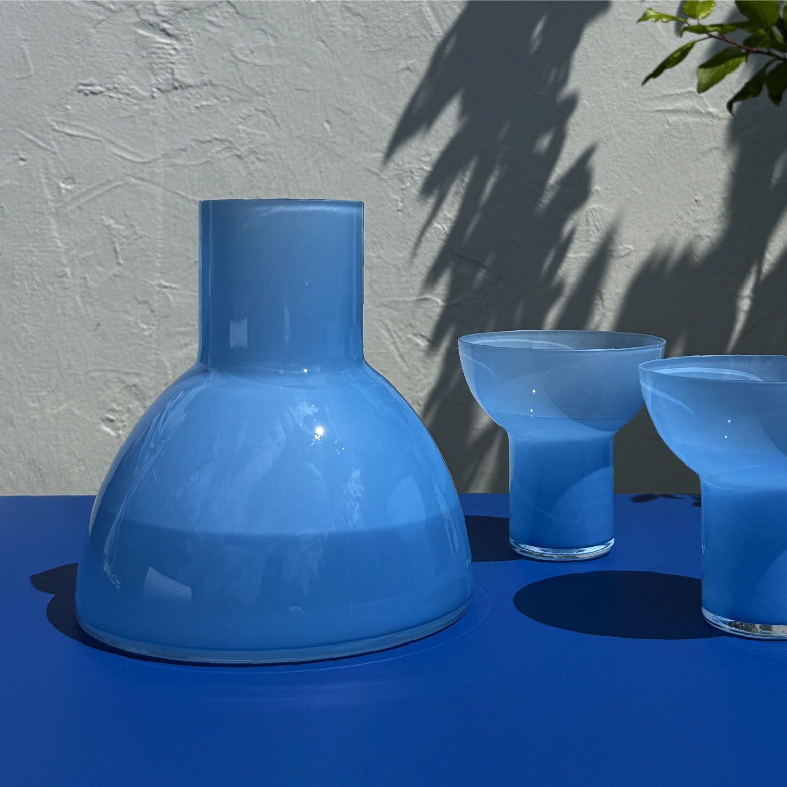 A light blue glass carafe with a wide rounded base and a short neck, shown alongside two matching blue plash glasses on a blue surface against a textured light wall with leaf shadows.