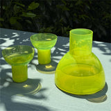 Three translucent lime green glassware pieces, including a large bulb-shaped carafe and two matching glasses, arranged on a light gray table outdoors with dappled sunlight and leaf shadows.