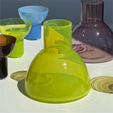 A translucent glass carafe in bright chartreuse yellow-green sits on a white surface, surrounded by other colored glass vessels including blue, purple, green, and amber tones under direct sunlight.