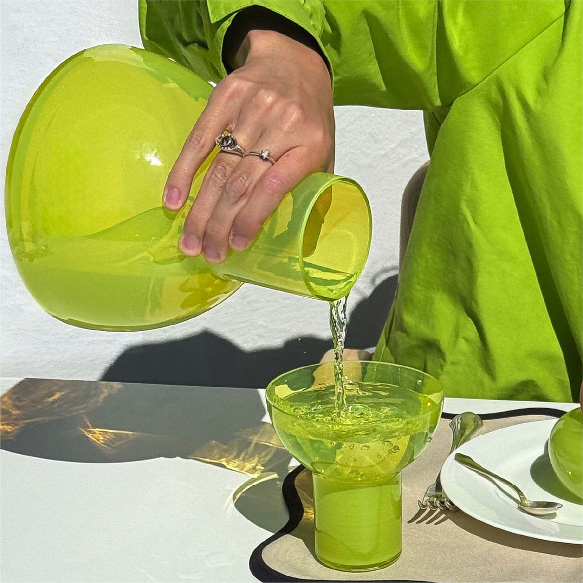 A person in a bright lime green outfit pours clear liquid from a translucent lime green carafe into a matching clear glass on a table.