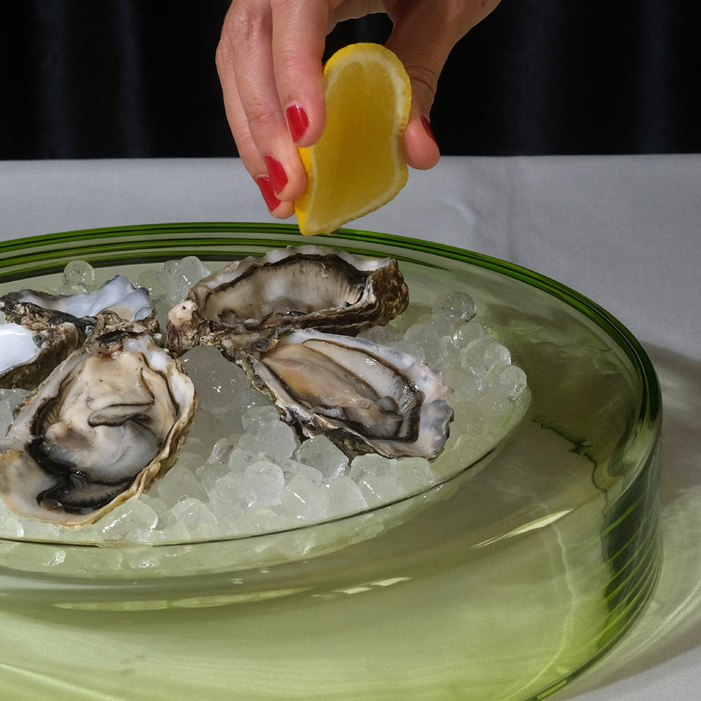 A close-up of a hand with red nail polish squeezing lemon juice onto several raw oysters resting on crushed ice in a lime wetbowl.