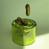 A high translucent lime-green wetbowl, containing a few medium sized pickles, with one pickle dropping into the bowl. Positioned on against a pale green background.