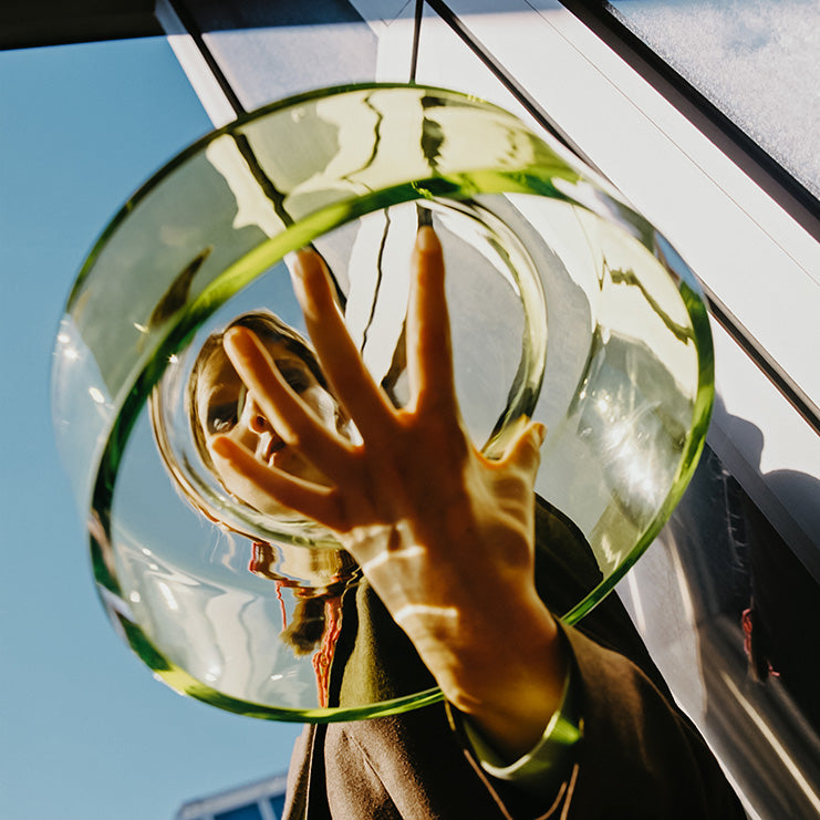 A person holds a large clear glass wetbowl in front of their face, with their hand reaching toward the glass. Strong sunlight creates dramatic shadows and reflections, and part of a modern building exterior and blue sky are visible in the background.