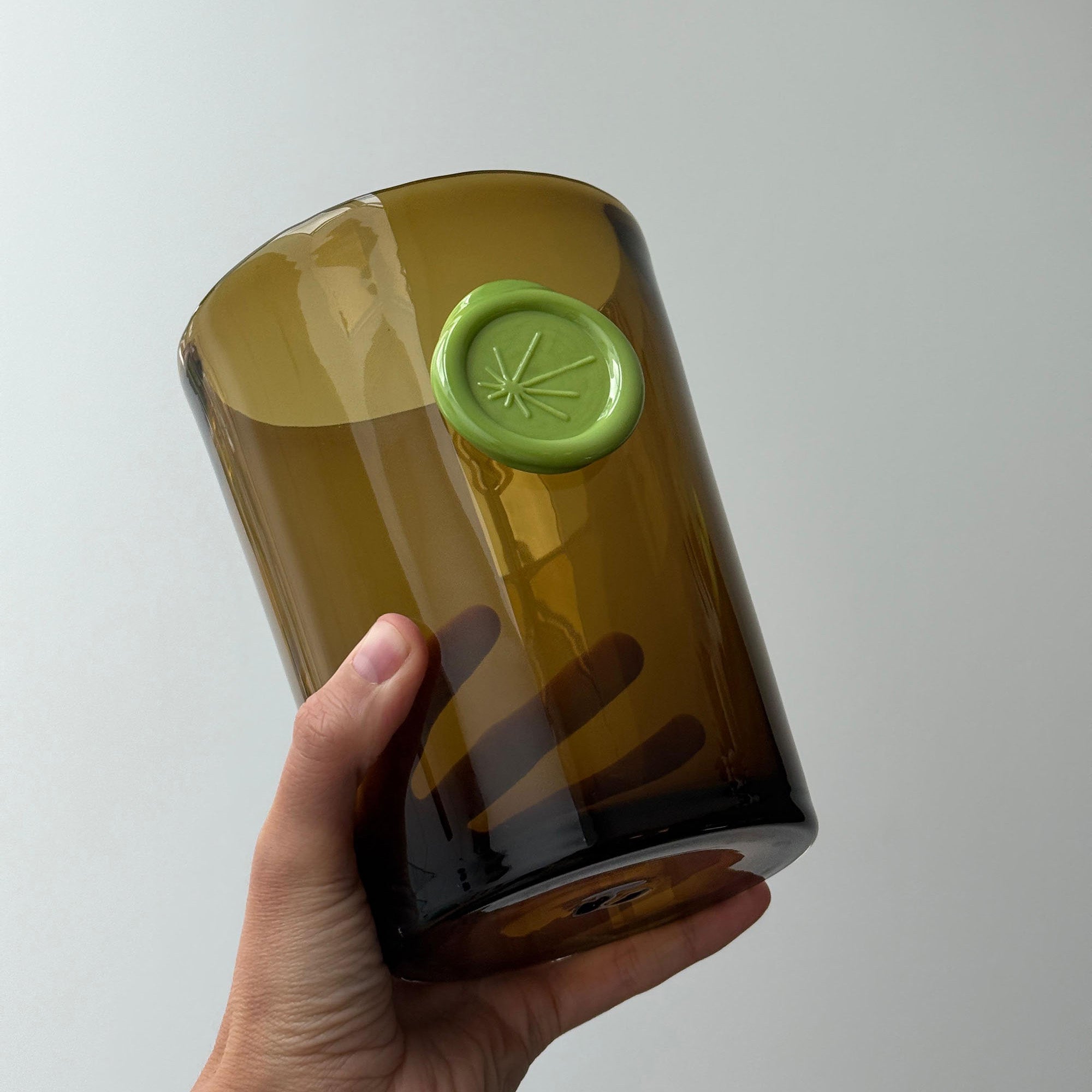 Hand holding a brown cylindrical vase with a green imprint on front, on a light gray background.