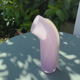 Video of the soft pink Nessie Carafe with a white strip on the back. Curved shape. On top of a table in a natural setting. The camera rotates around the carafe.